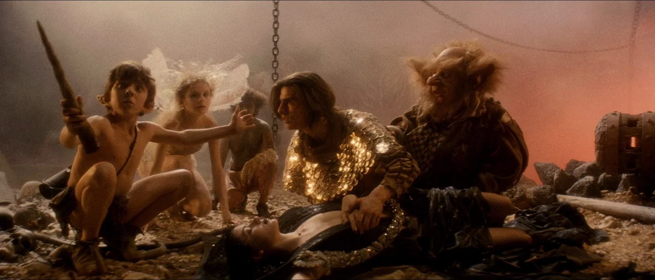 Legend (1985) movie still: medium — A dimly lit rocky cavern floor glows with a warm, fiery, orange-tinted background haze. A diverse group of…