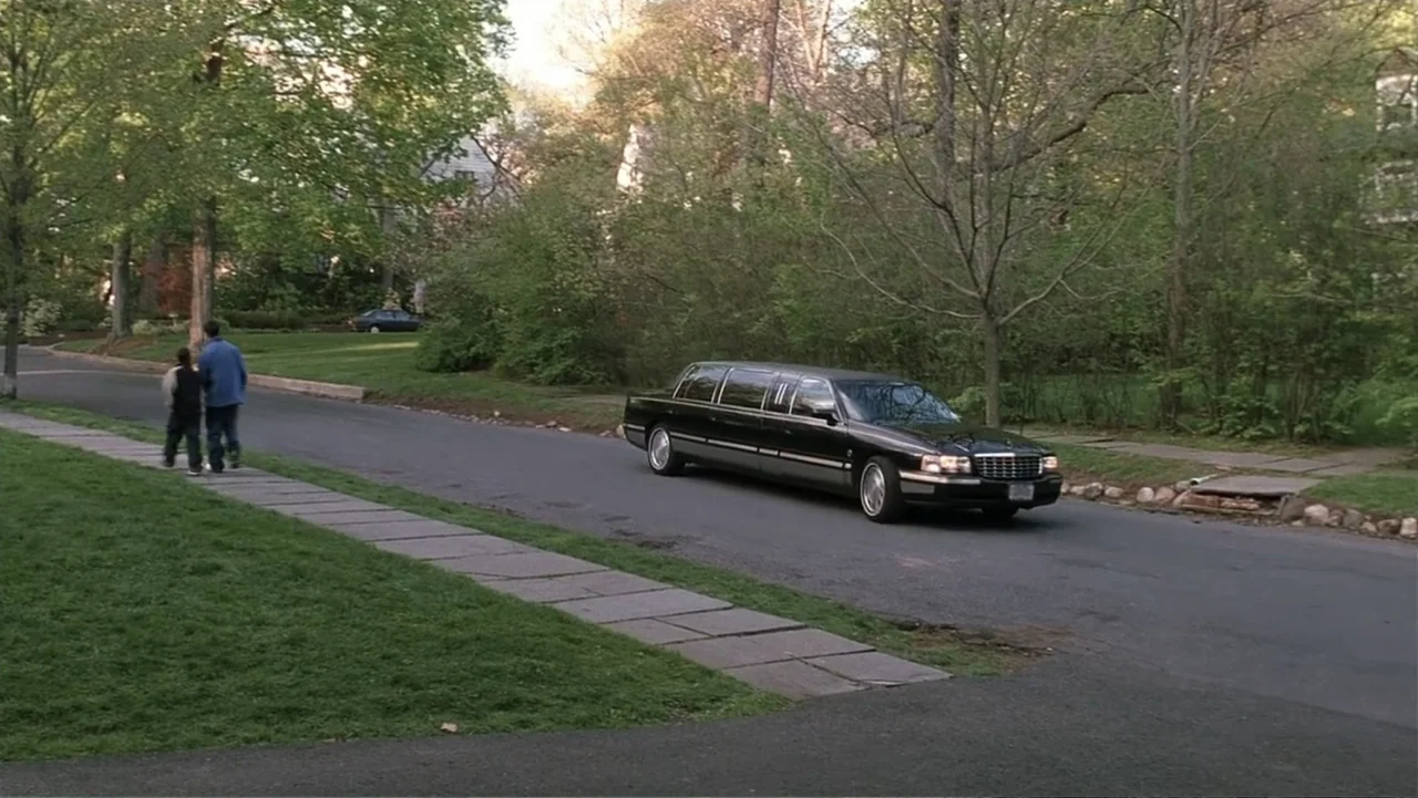 Analyze That (2002) movie still: wide — A quiet residential street lined with mature trees and green grass during the day. A black limousine drives…