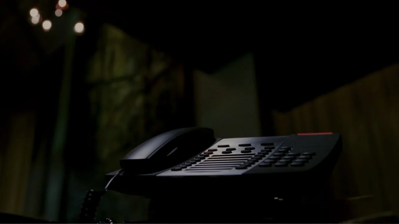 When a Stranger Calls (2006) movie still: closeup — A dark office interior features a black telephone sitting on a desk surface. Small bokeh…
