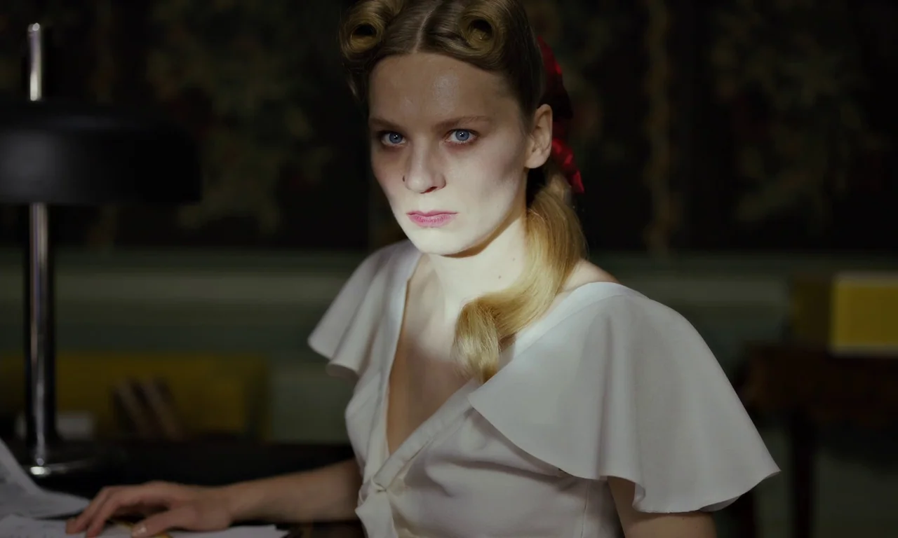 Bloodsuckers (2021) movie still: medium — A dimly lit room features a woman seated at a desk with a lamp. The woman stares at the camera while the…