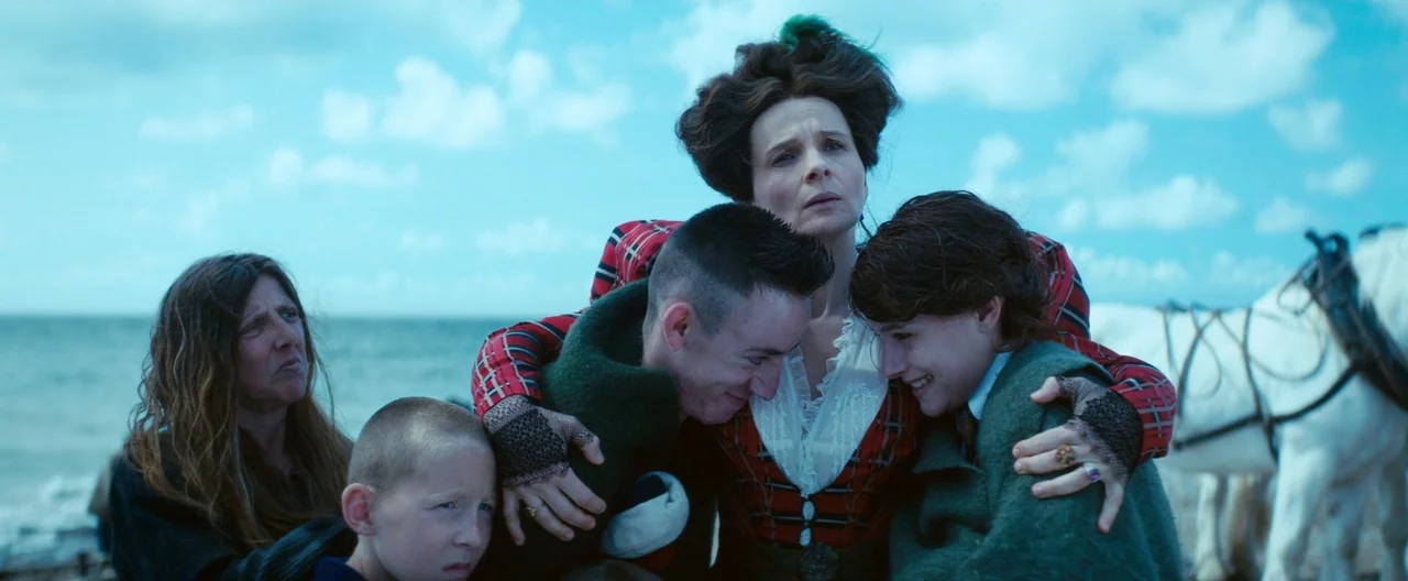 Slack Bay (2016) movie still: medium — A group of people stand together on a sandy beach under a bright blue sky. A woman in a plaid jacket holds…