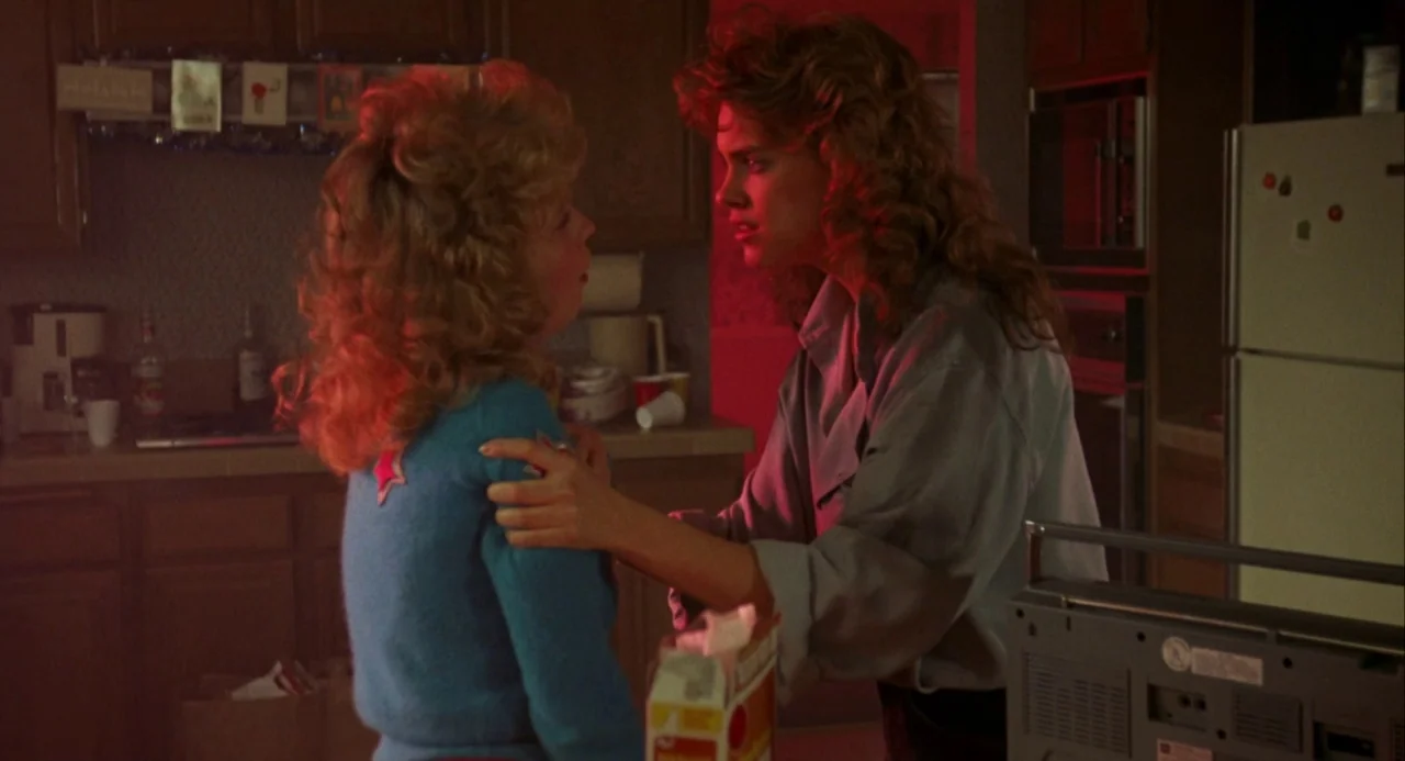 Night of the Comet movie still: medium — A residential kitchen features dark brown cabinetry, a refrigerator, and a small boombox. One woman firmly…