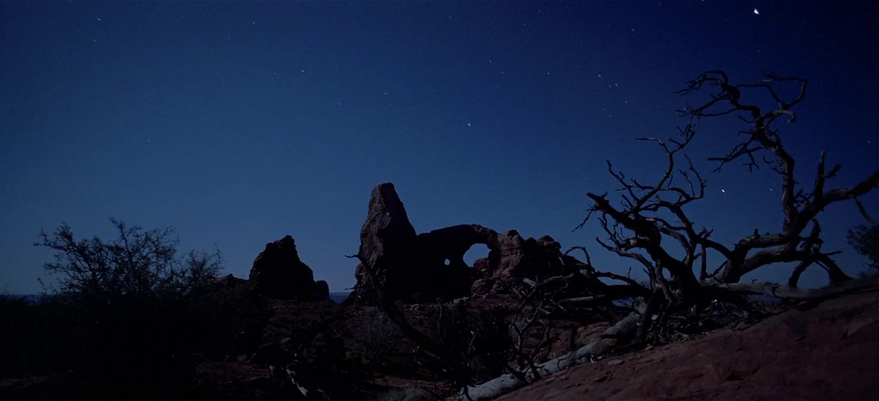 Baraka (1992) movie still: establishing — A vast desert landscape features large sandstone rock arches silhouetted against a night sky. A gnarled…