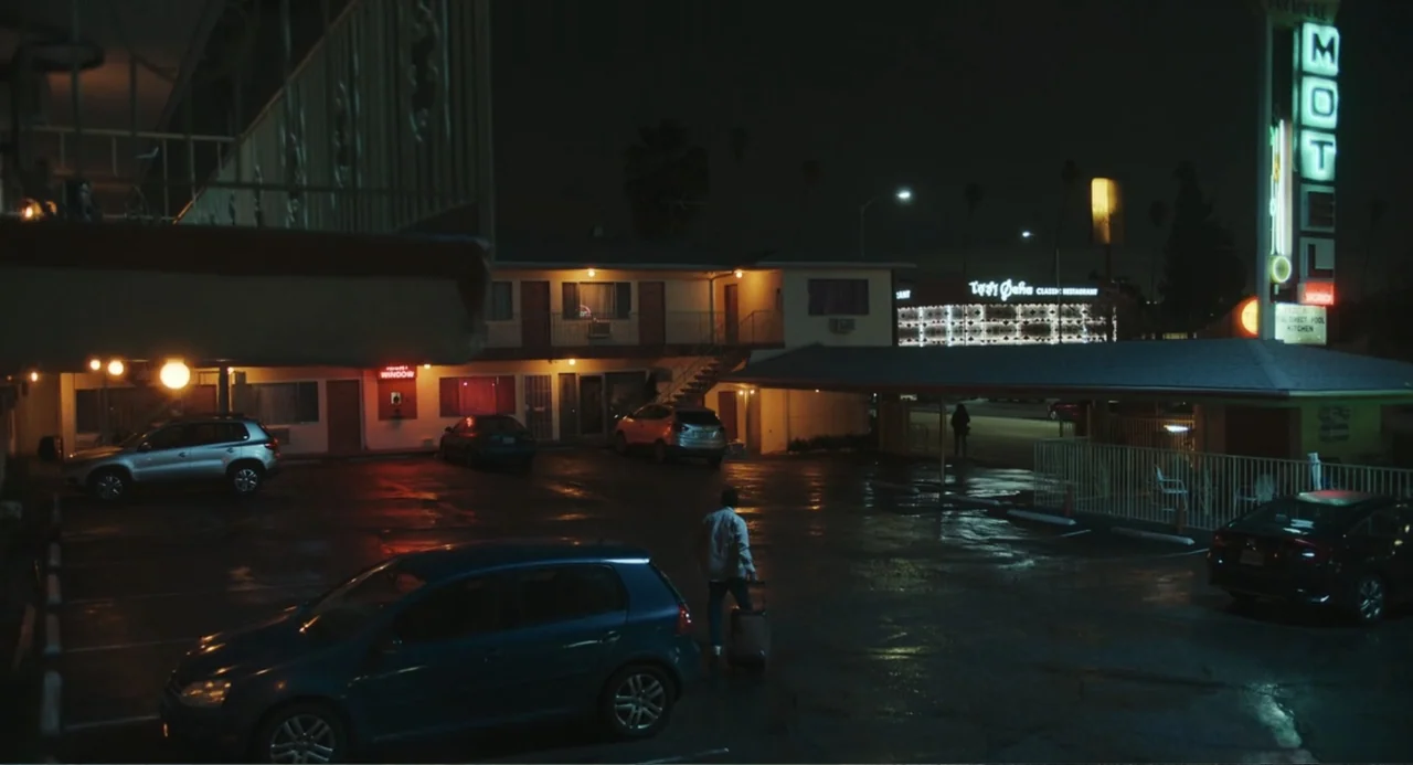 Good Fortune (2025) movie still: wide — A wet asphalt motel parking lot at night with several parked cars under illumination. A lone individual…