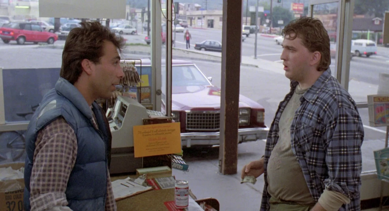 River’s Edge (1986) movie still: medium — A bright convenience store interior overlooks a street with parked cars through glass windows. Two men…