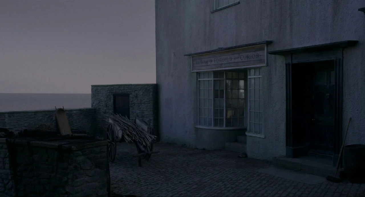 Ammonite (2020) movie still: establishing — A stone building with a small shopfront faces a calm, dark sea at twilight. A wooden cart loaded with…