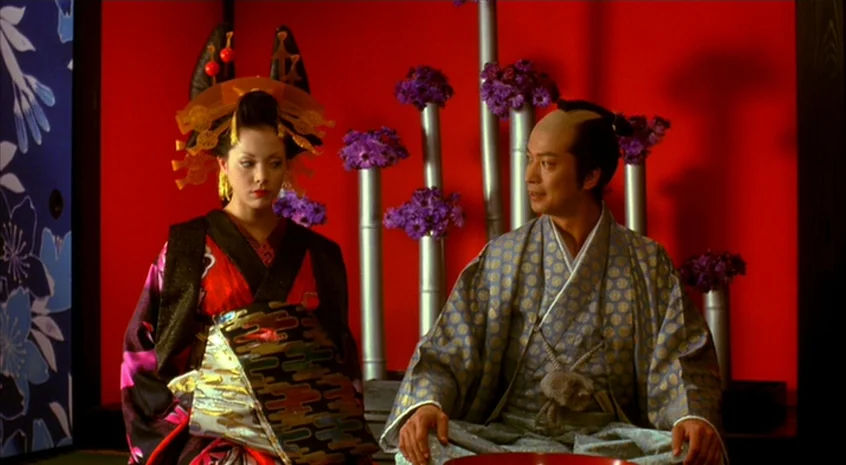 Sakuran (2006) movie still: medium — An interior space features a bold red wall decorated with vertical metallic tubes. A woman and a man in…