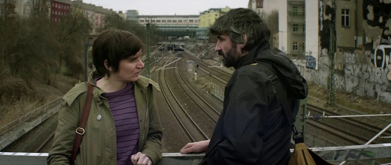 Silence (2012) movie still: medium — A pedestrian bridge overlooks railway tracks running through a dense, urban environment with buildings. Two…