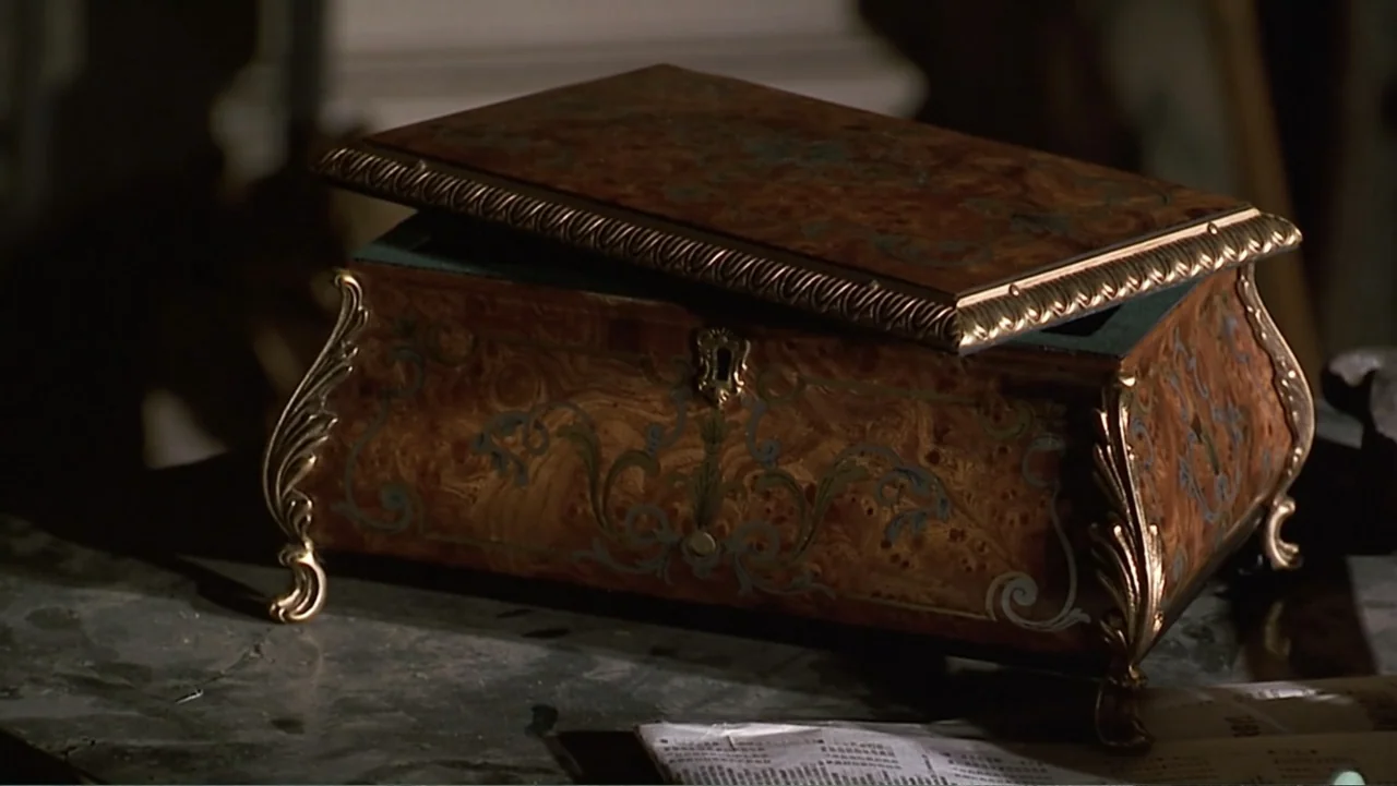 Matilda (1996) movie still: closeup — An ornate wooden jewelry box with inlaid patterns sits on a dark marble tabletop. The lid is raised slightly,…