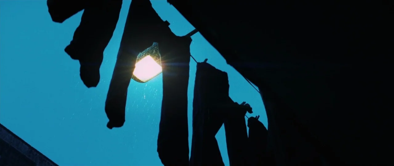 Biutiful (2010) movie still: wide — Dark silhouettes of clothing hang from a line against a deep blue night sky. A bright streetlamp illuminates the…