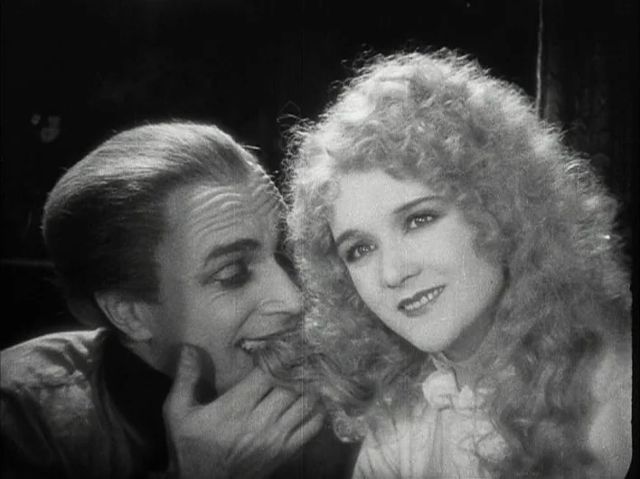 The Man Who Laughs (1928) movie still: closeup — A dark, minimally lit room with a neutral background. A man leans in closely to a woman, playfully…