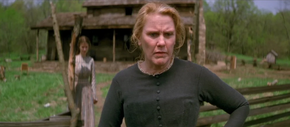 Ride With The Devil movie still: medium — A rural farm with a wooden cabin sits in a green field. A mature woman stands in the foreground while a…