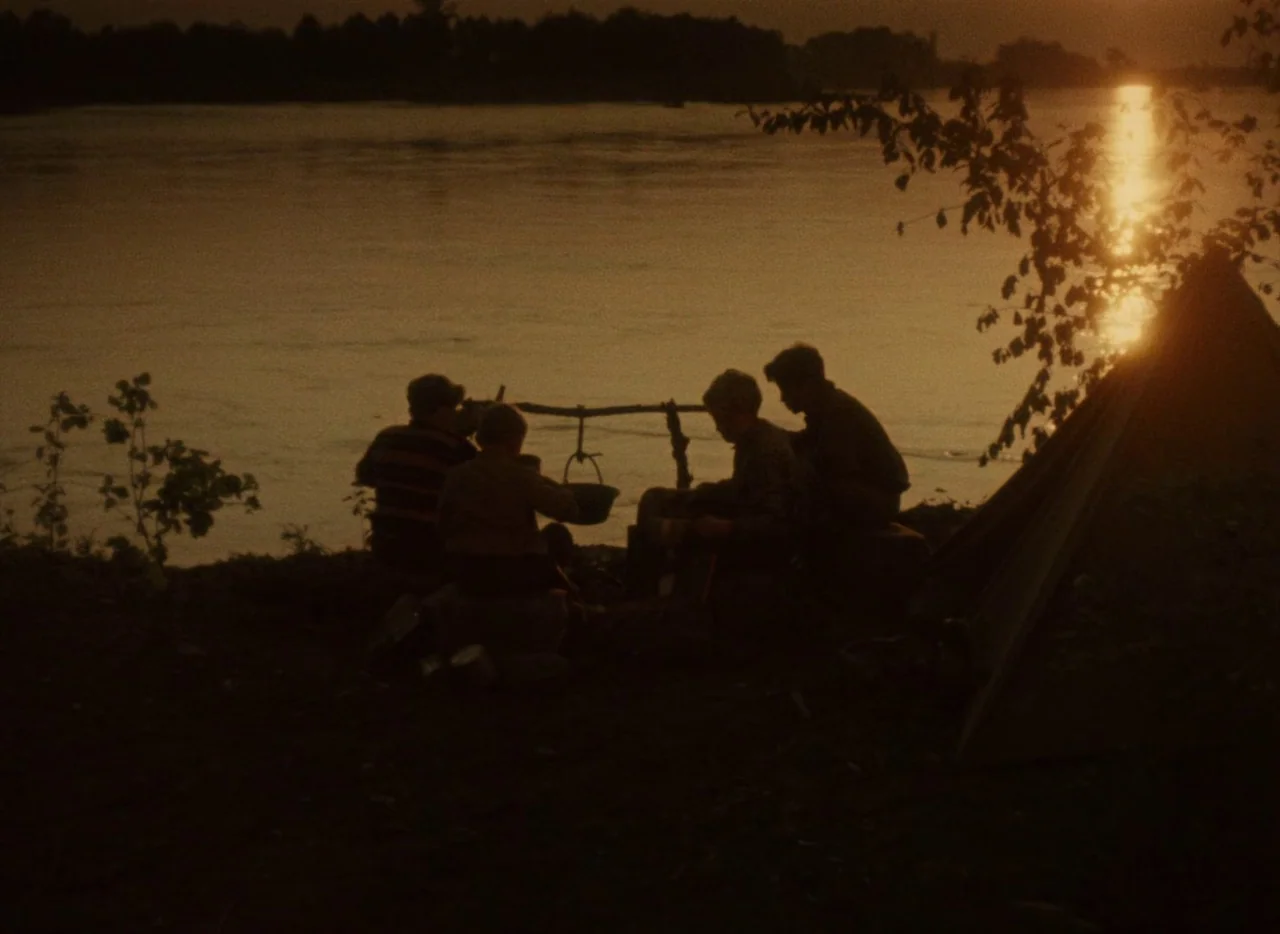 A Journey to the Beginning of Time (1955) movie still: wide — Four figures sit in silhouette on a riverbank during the golden hour sunset. A small…