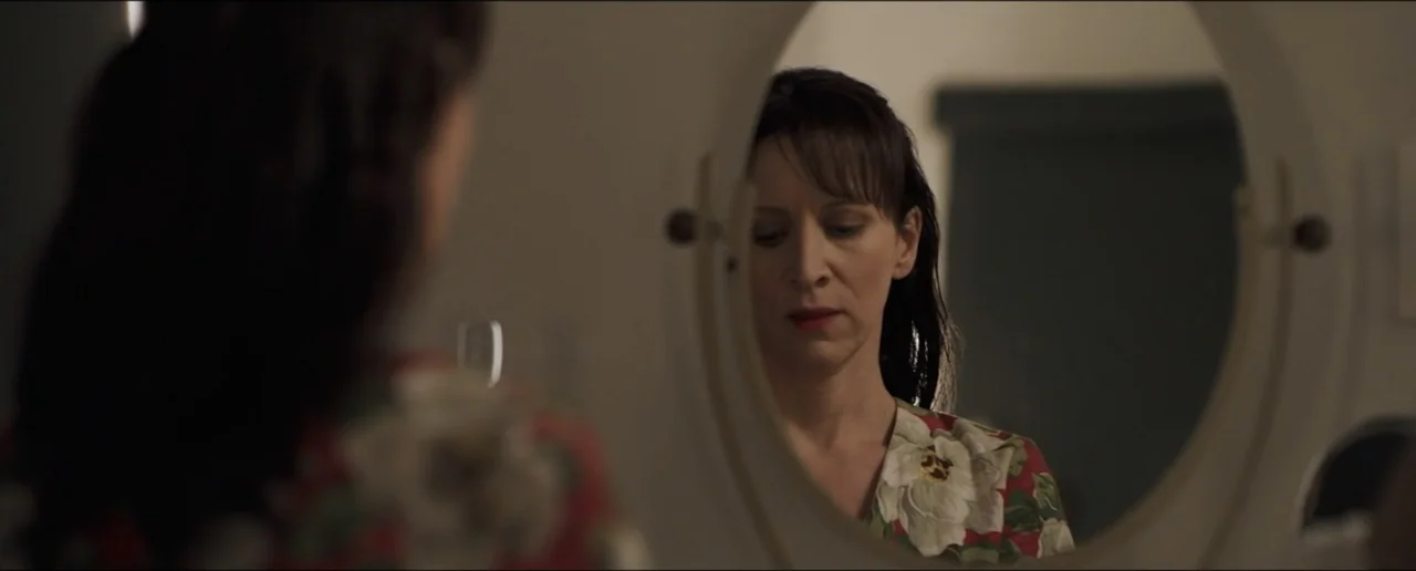 Hostages (2017) movie still: medium — A dimly lit room features a woman reflected in a simple oval vanity mirror. The subject sits facing the mirror…