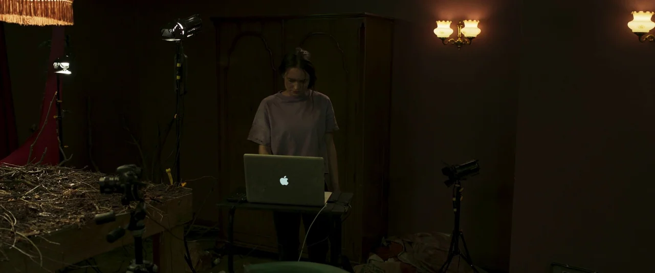 Stopmotion (2024) movie still: medium — A dimly lit production space featuring a laptop on a desk and studio equipment. A young woman stands behind…