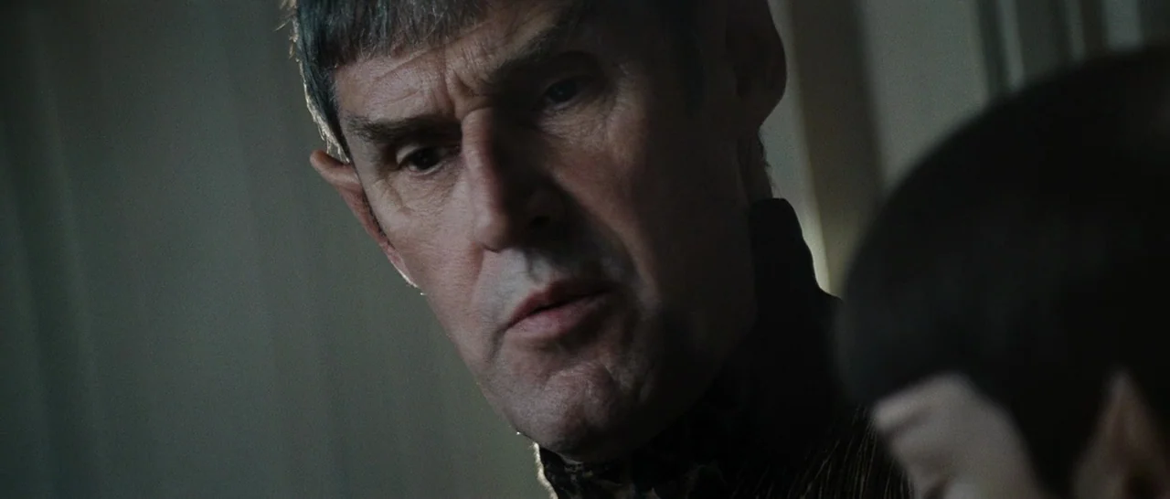 Star Trek (2009) movie still: closeup — A dimly lit interior space with minimal architectural detail visible in the shadows. A Vulcan man with…