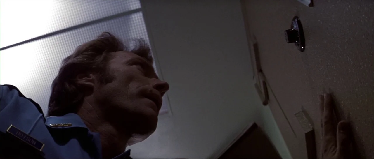 Thunderbolt & Lightfoot (1974) movie still: medium — A dimly lit indoor corridor with an exposed fluorescent ceiling panel above. A man looks up at…