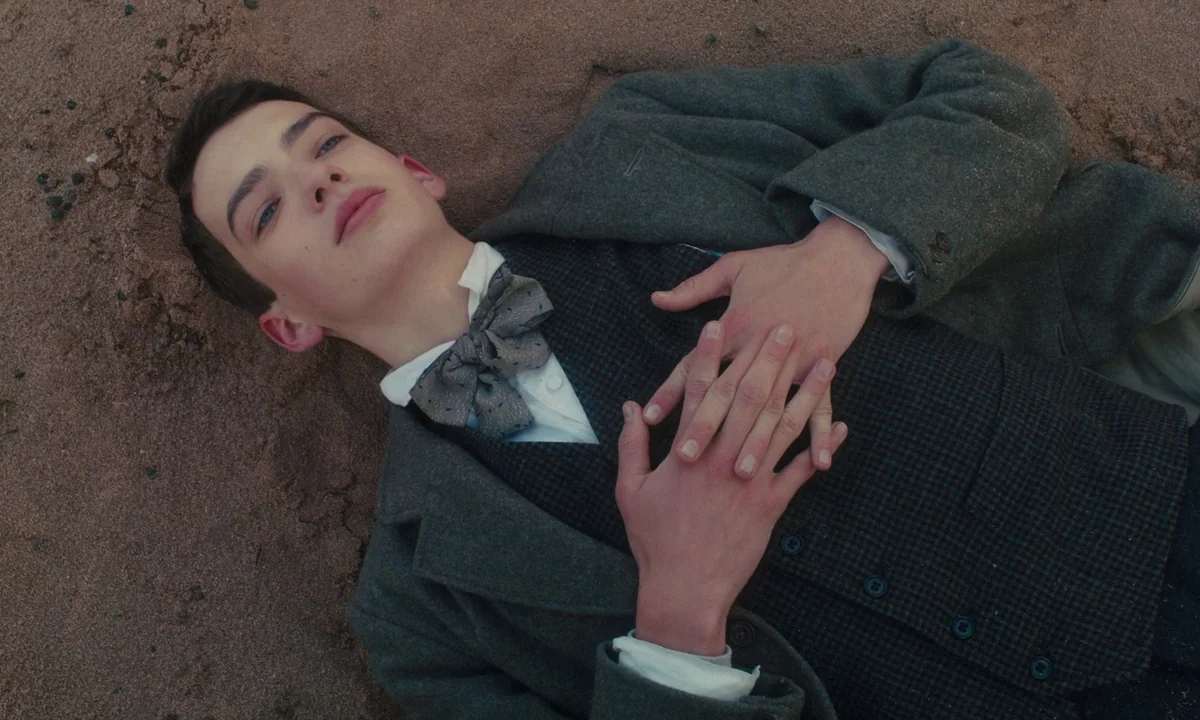 Slow West (2015) movie still: medium — A young man lies flat on a sandy surface. He wears formal period clothing and rests his hands over his chest.
