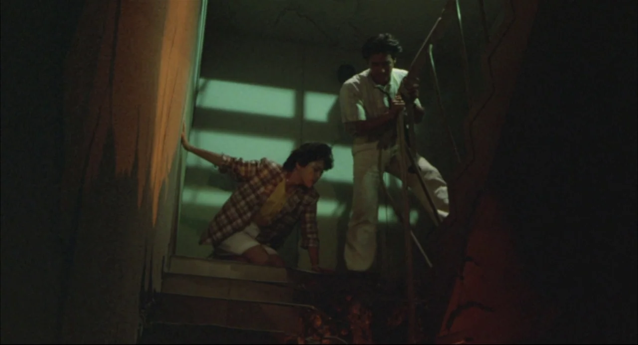 The Return of Godzilla (1984) movie still: fullbody — A dimly lit industrial staircase with green wall paint and metal railings. A woman crouches on…
