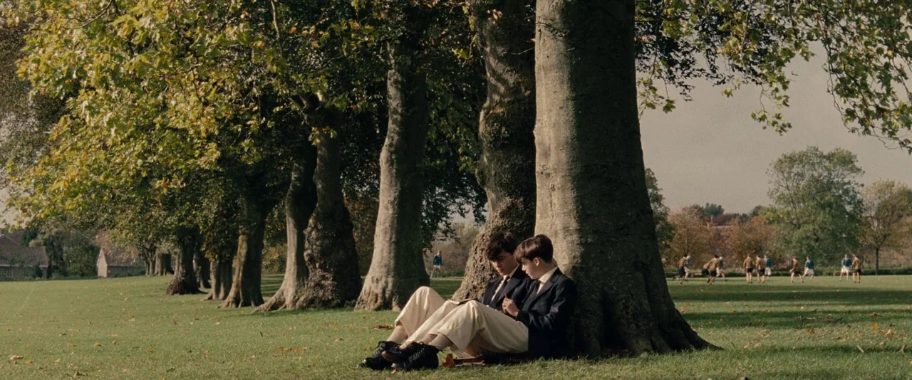 The Imitation Game (2014) movie still: wide — Two boys sit at the base of a massive tree in a grassy meadow. In the distance, other students play on…