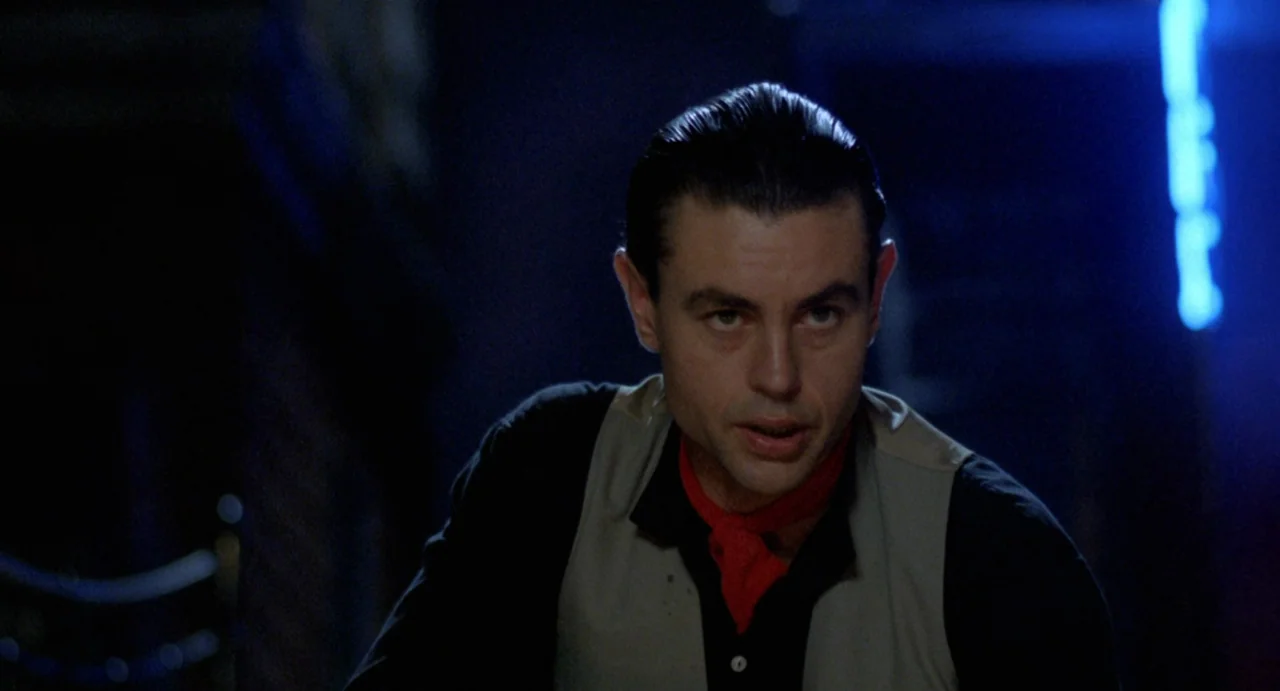 StageFright (1987) movie still: medium — A dark space at night with a blue neon light in the background. A man stands wearing a cream vest and red…