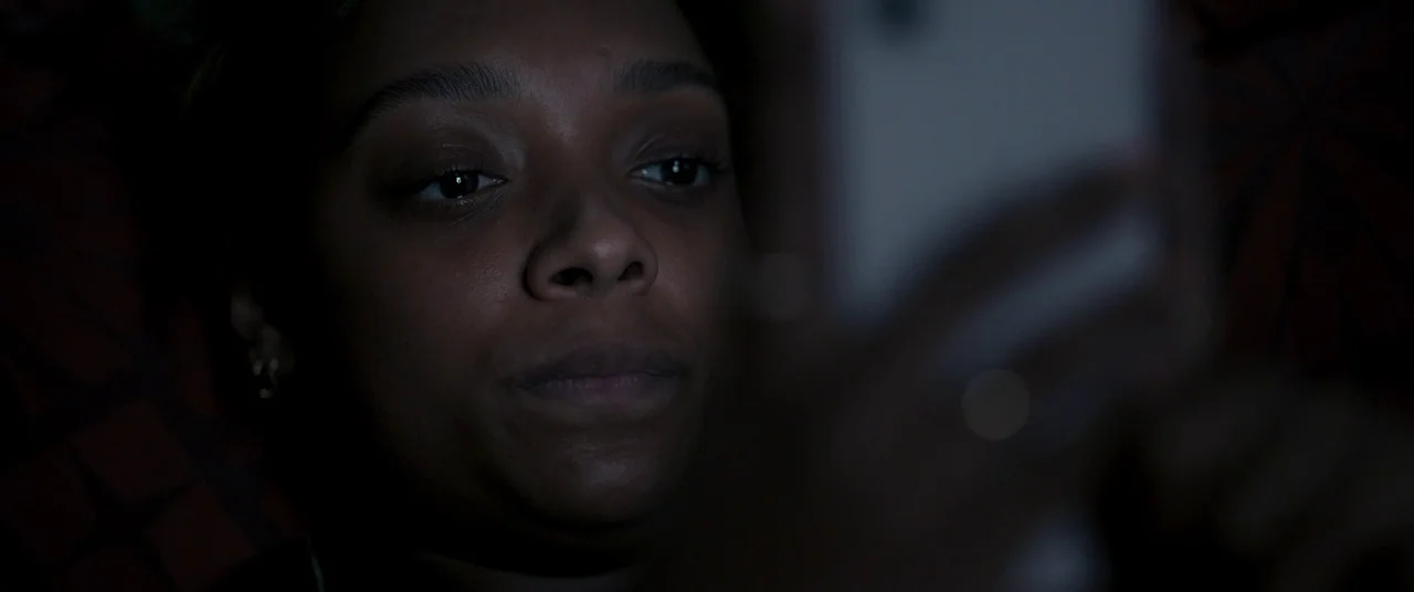 Talk To Me (2022) movie still: closeup — A woman stares into the light of her phone in a pitch black room. The soft illumination highlights her…