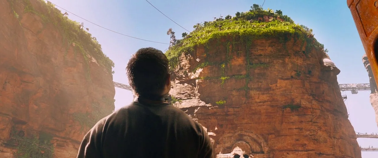 Mad Max: Fury Road (2015) movie still: medium — A massive vertical rock formation with dense vegetation stands within a deep arid canyon. A man seen…