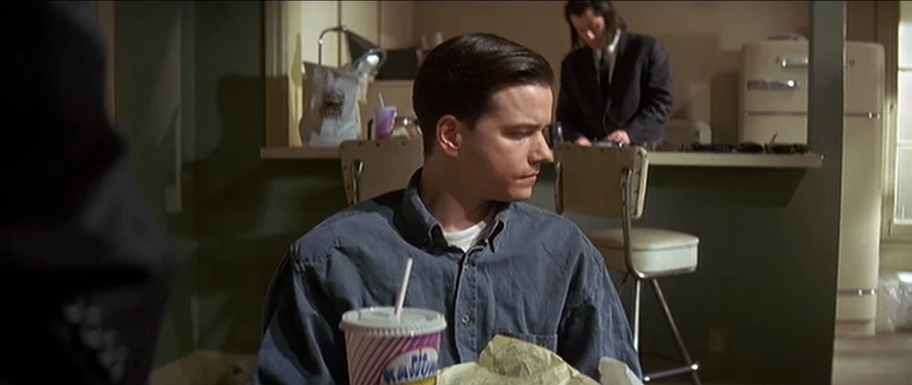 Pulp Fiction (1994) movie still: medium — A sparse room features a counter with a stool and a vintage refrigerator. One man sits in the foreground…