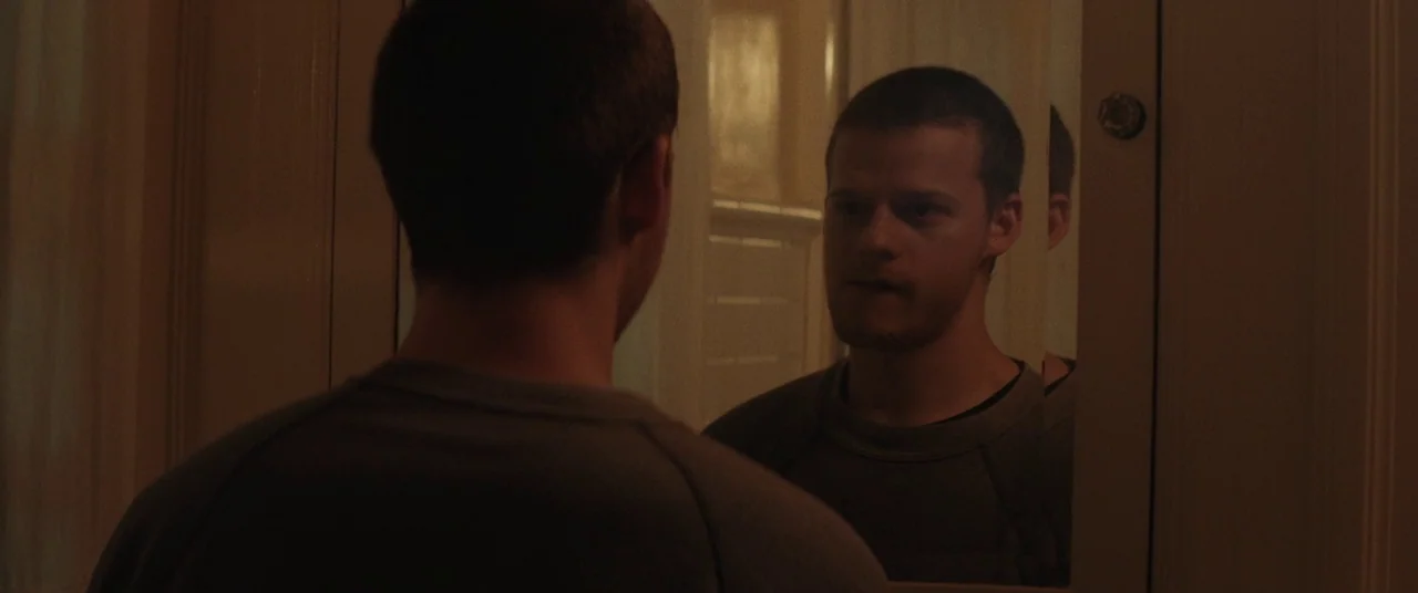 Honey Boy (2019) movie still: medium — A dimly lit room with a reflective glass surface and beige wall panels. A young man stands facing his own…
