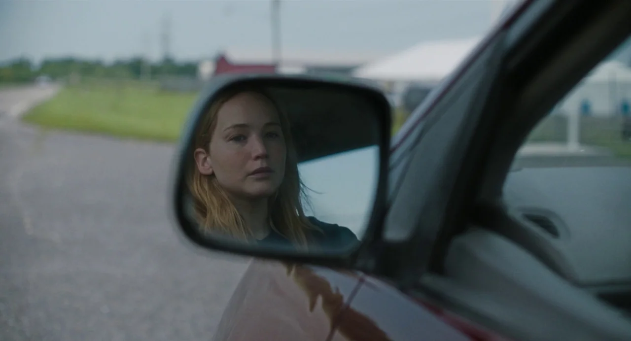 Causeway movie still: closeup — A view focused on the side mirror of a vehicle parked on a road. A young woman is clearly visible reflected in the…