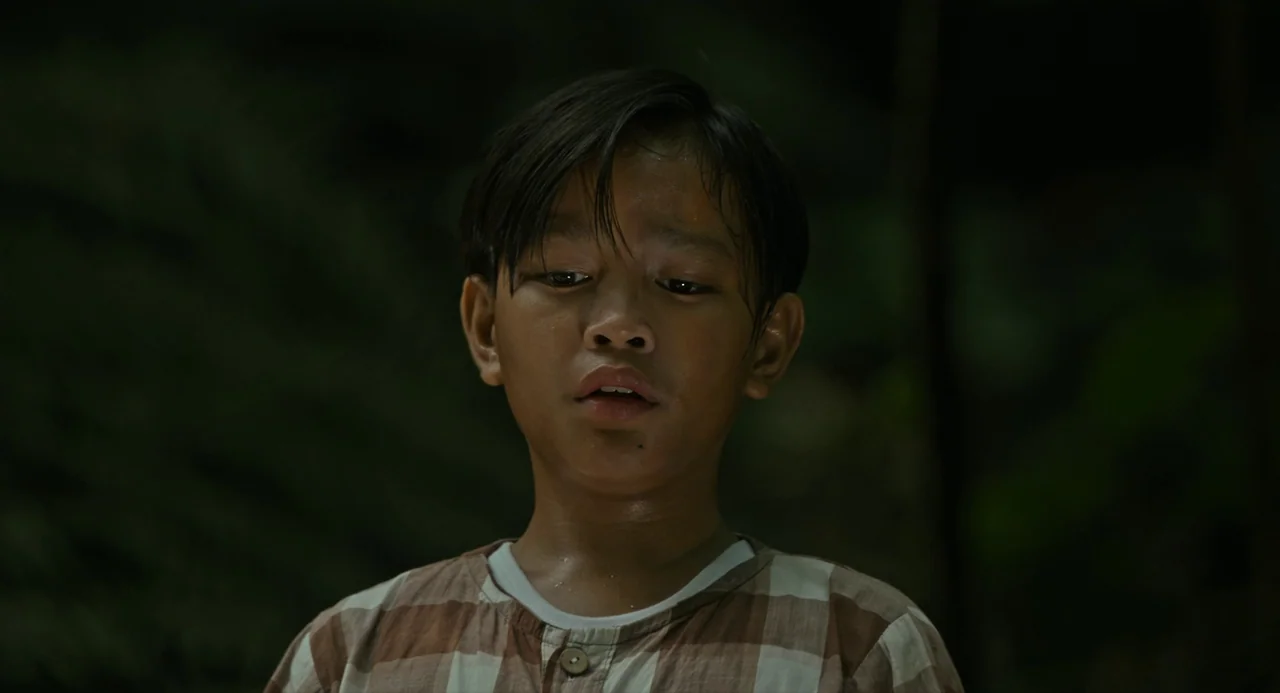 In My Mother’s Skin (2023) movie still: closeup — A young boy stands in a dark, outdoor setting at night. He looks downward with a soft, neutral…