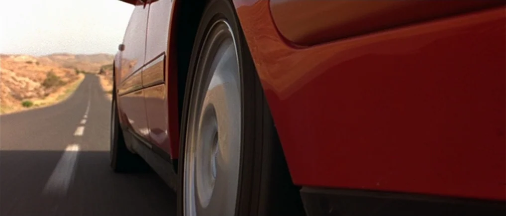 Sexy Beast (2000) movie still: closeup — A bright red car drives along an asphalt road through an arid desert environment. The camera is mounted low…