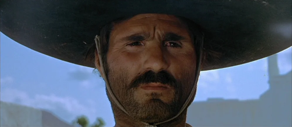 For A Few Dollars More (1965) movie still: closeup — An outdoor town setting with pale buildings and a bright blue sky. A man in a dark sombrero…