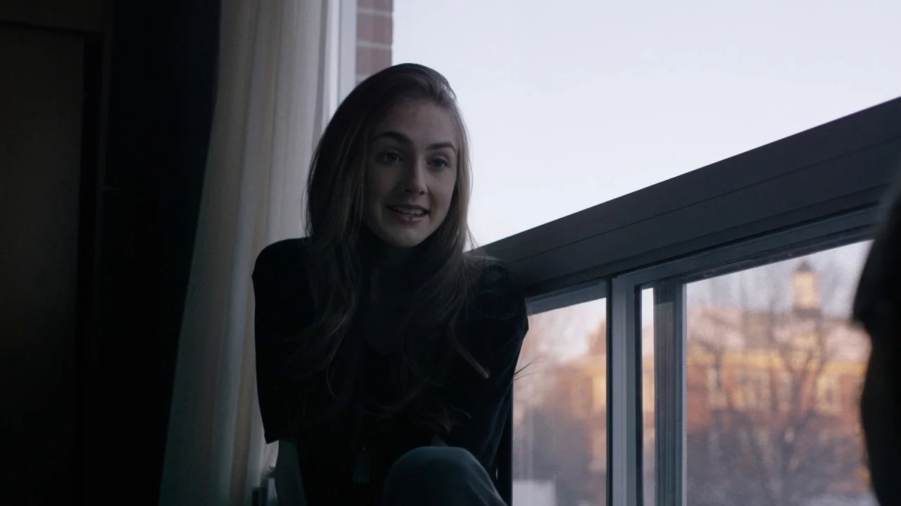 The Blackcoat’s Daughter (2015) movie still: medium — A woman sits next to a large window with a blurry outdoor background visible. She looks toward…