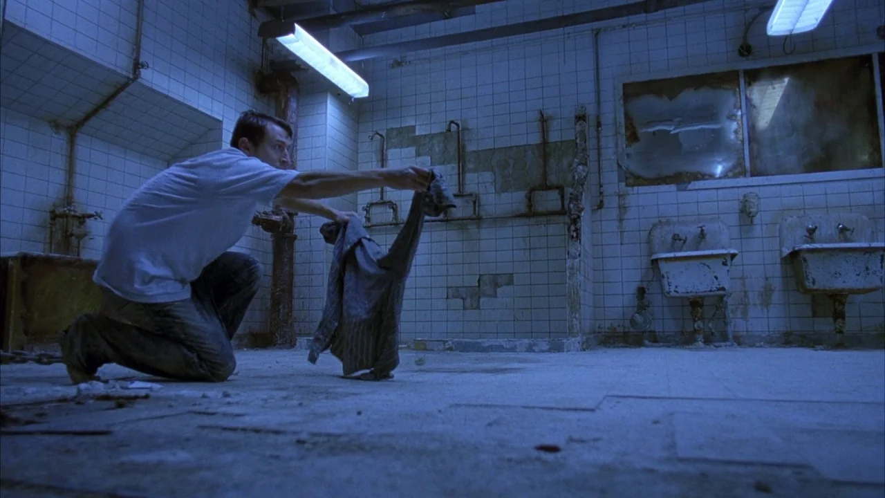Saw movie still: fullbody — A decrepit, tiled room features exposed plumbing pipes and two dirty washbasins on walls. The man kneels on the gritty…
