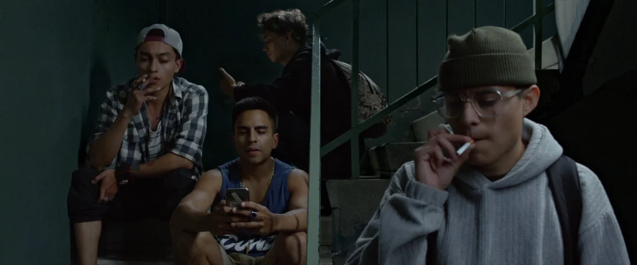 Like or Die (2023) movie still: medium — A dimly lit concrete stairwell with shadows filling the background space. Four young men sit on the steps,…