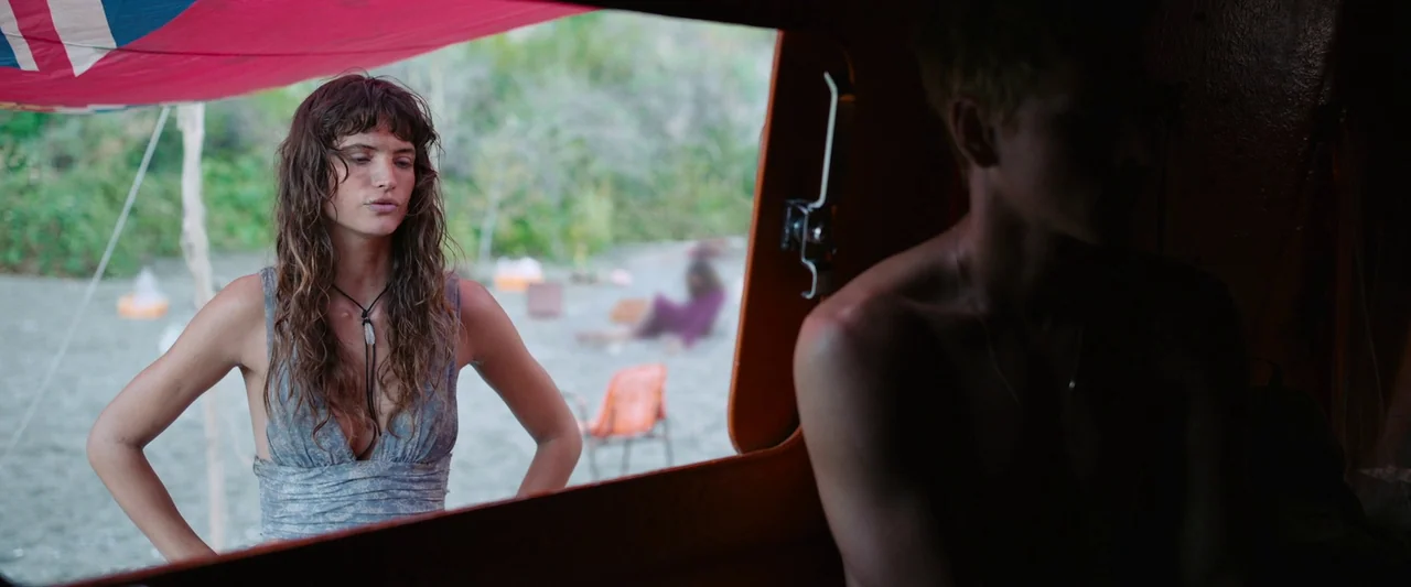 Triangle of Sadness (2022) movie still: medium — A woman stands outside an open vehicle frame on a beach with sparse foliage. A second person is…