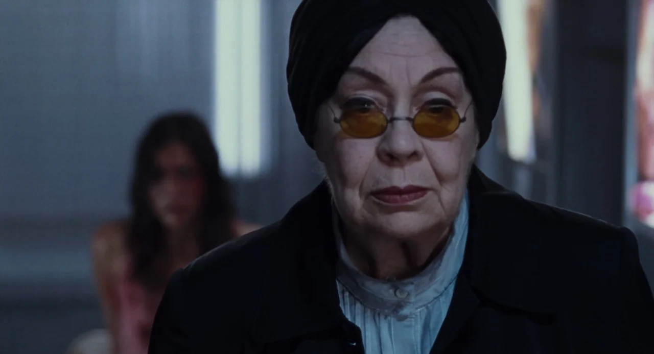 Martyrs (2008) movie still: closeup — An elderly woman stands in the foreground inside a dark, nondescript interior space. A second out-of-focus…
