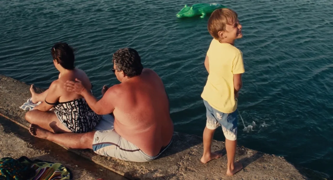 Ava (2017) movie still: medium — A man and woman sit on a stone pier next to calm blue water. A young boy stands nearby and urinates into the water…