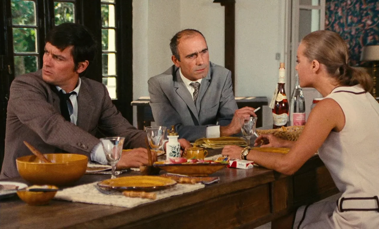 The Swimming Pool (1969) movie still: medium — A rustic dining room features a long wooden table set with plates and glasses. Two men and one woman…