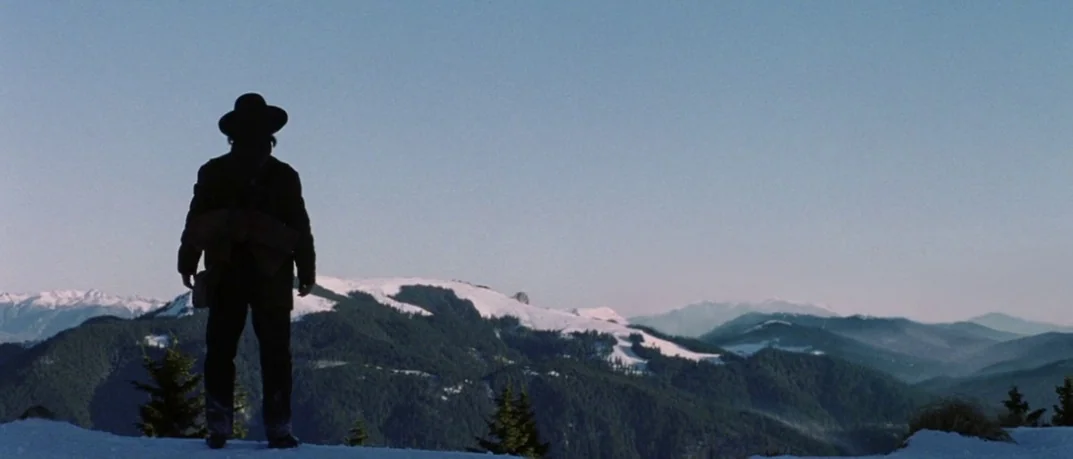 Cold Mountain (2003) movie still: wide — A snow-covered mountain ridgeline overlooks a series of forested mountains in the distance. A man in a dark…