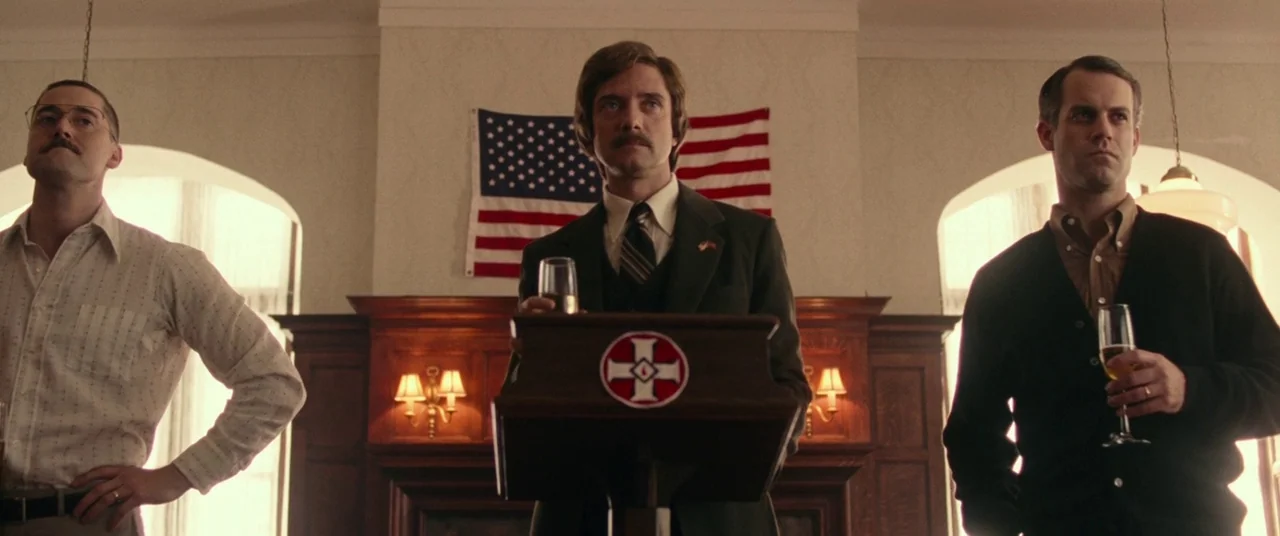 BlacKkKlansman (2018) movie still: medium — Three men stand before a podium in a wood-paneled room. An American flag hangs on the wall behind them…