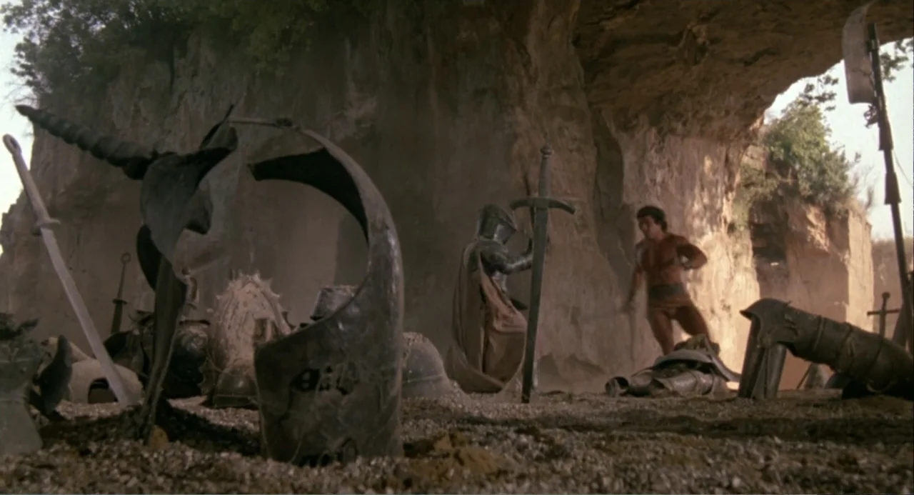Hercules (1983) movie still: wide — A rocky cavern floor is littered with various discarded helmets, swords, and plate armor pieces. A muscular man…