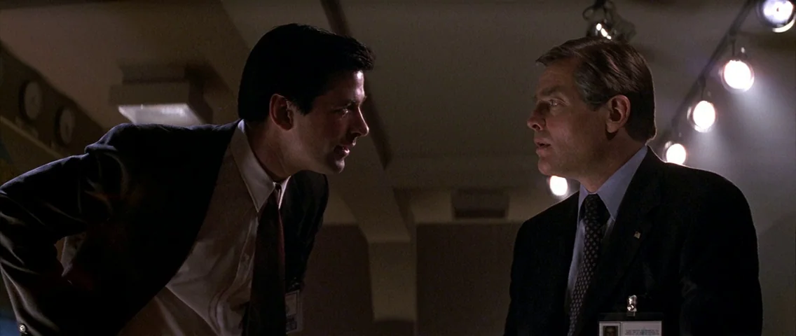 The Hunt for Red October (1990) movie still: medium — A dimly lit professional office with track lights mounted on the ceiling above. Two men in…