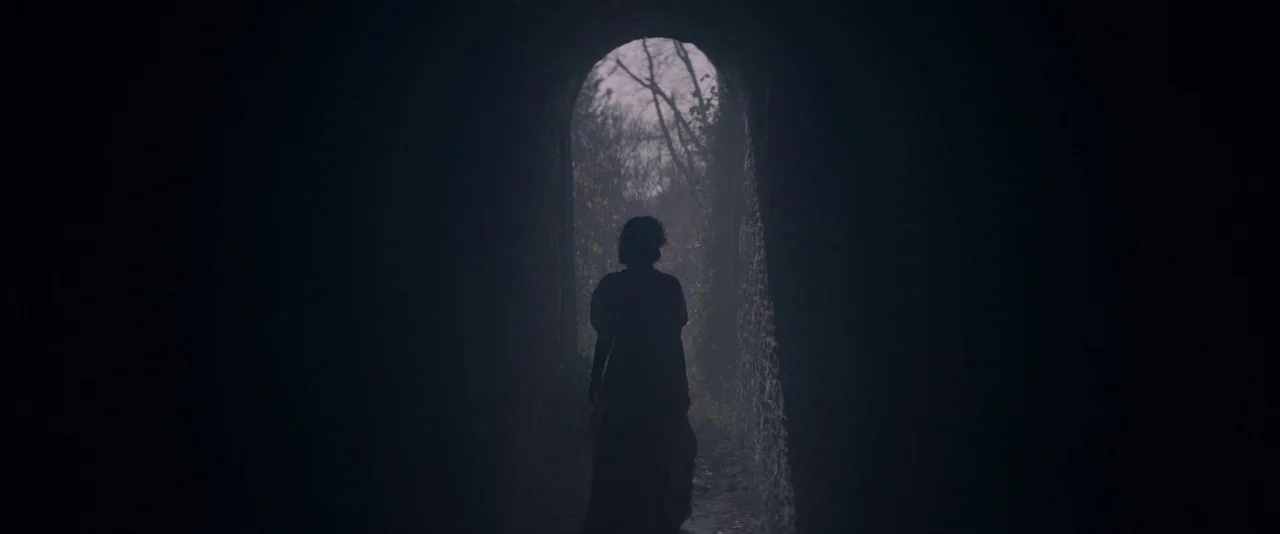 The Dancer movie still: fullbody — A dark, narrow stone archway frames a view of a misty, leafless winter woodland. A single woman in a period-style…