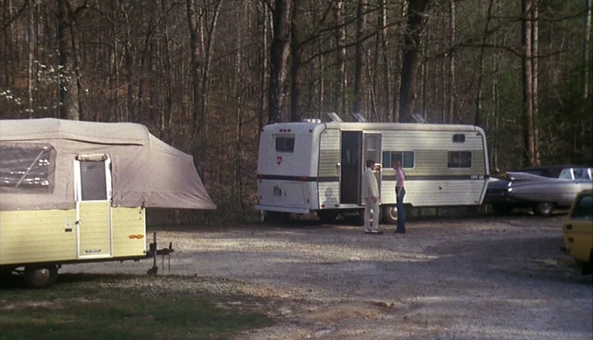 Cockfighter (1974) movie still: wide — A gravel-surfaced campsite is surrounded by a dense woodland of tall pine trees. A man and a woman stand…