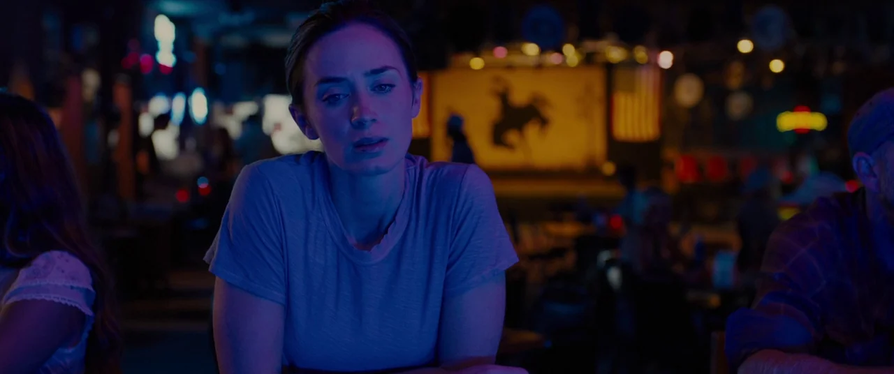 Sicario (2015) movie still: medium — A dark dimly lit bar environment with blurred patrons and ambient neon signage. A woman in a plain t-shirt sits…