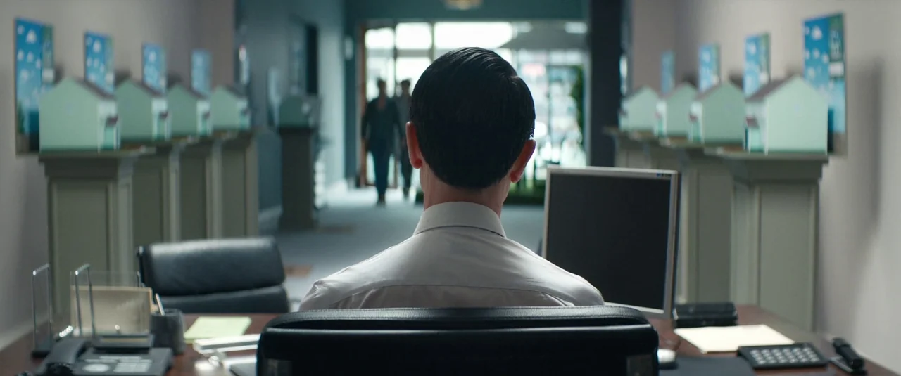 Vivarium (2019) movie still: medium — An office interior features rows of model houses on pedestals lining a long hallway. A man in a white shirt…