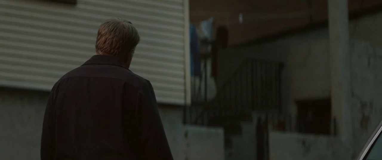 God’s Pocket (2014) movie still: medium — A residential exterior features a light-colored siding building and a dark concrete stairway. A man in a…