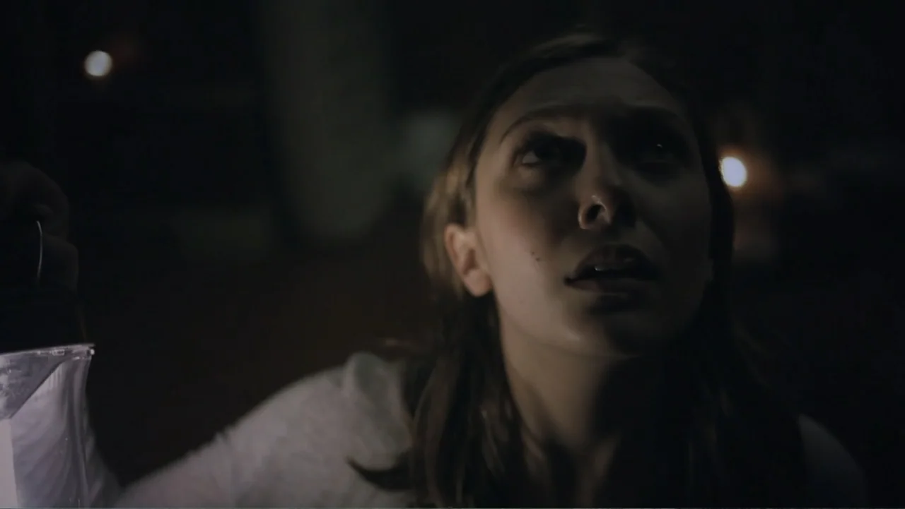 Silent House (2011) movie still: closeup — A dark, dimly lit indoor space with shadows obscuring the surroundings. A young woman in a grey shirt…